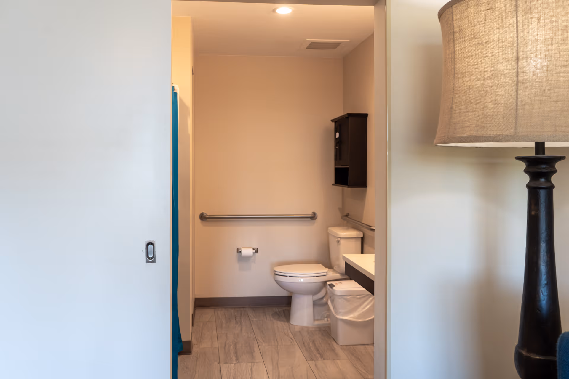 View into a bathroom with a white toilet, a grab bar on the wall, a toilet paper holder, a small black wall cabinet, and a trash can. The bathroom has light-colored walls and tiled flooring. A lamp with a beige shade is partially visible on the right side outside the bathroom.