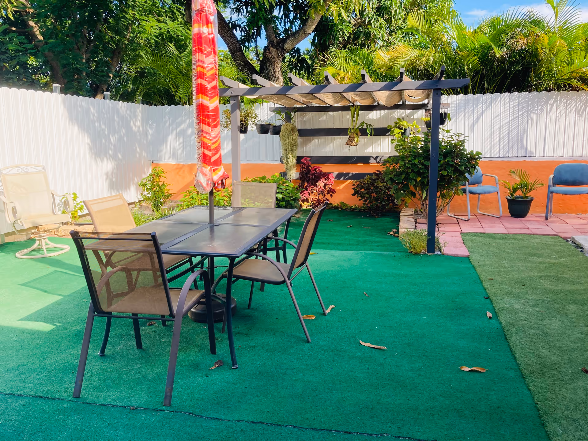 Outdoor patio area with a table and four chairs, a closed red umbrella in the center of the table, a pergola with hanging plants, green artificial turf flooring, and various plants and bushes along a white fence.