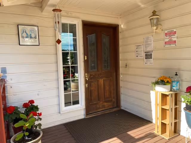 Wooden front door with decorative glass panels on a porch with beige siding walls. A tall narrow window is to the left of the door, and a brown doormat lies in front. There are potted plants with red and yellow flowers on either side, a wooden crate holding a white planter with yellow flowers, a hand sanitizer dispenser, a wall-mounted lantern light, a wind chime, and several posted notices on the wall.