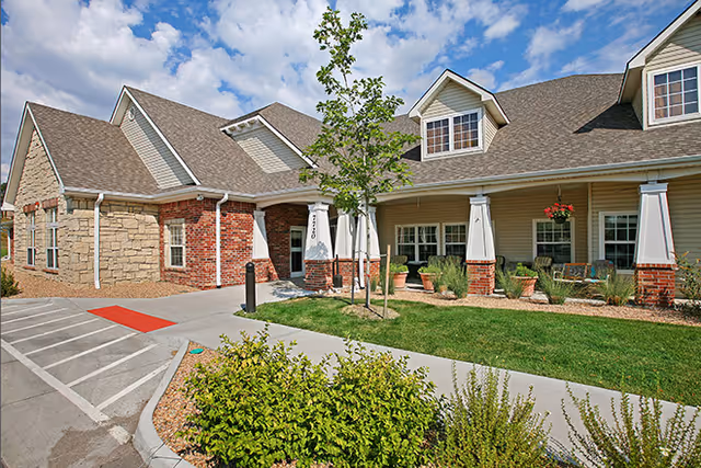 Exterior view of a senior living facility building with a combination of brick and stone walls, a covered entrance supported by white columns with brick bases, a small tree and landscaped greenery in front, and a partly cloudy sky overhead.