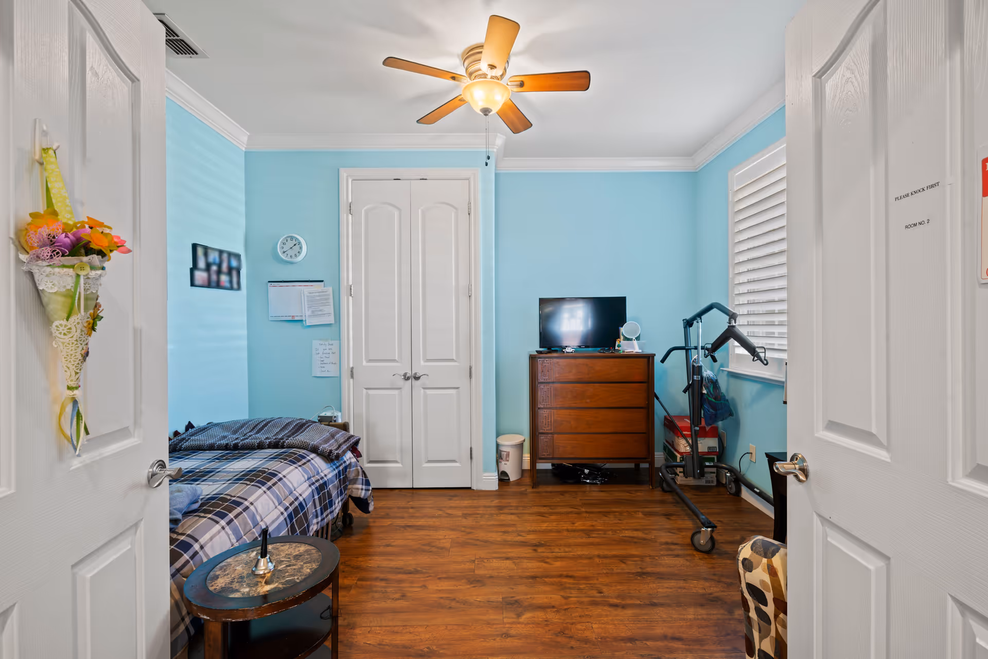 A small bedroom with light blue walls and wooden floor. The room contains a single bed with plaid bedding on the left, a wooden dresser with a small TV on top against the far wall, and a medical lift device near the window on the right. There is a ceiling fan with a light fixture above, a round side table next to the bed, and double white closet doors in the center of the far wall. Two white doors are partially open, one on each side of the image, with a decorative flower cone hanging on the left door and a sign on the right door.