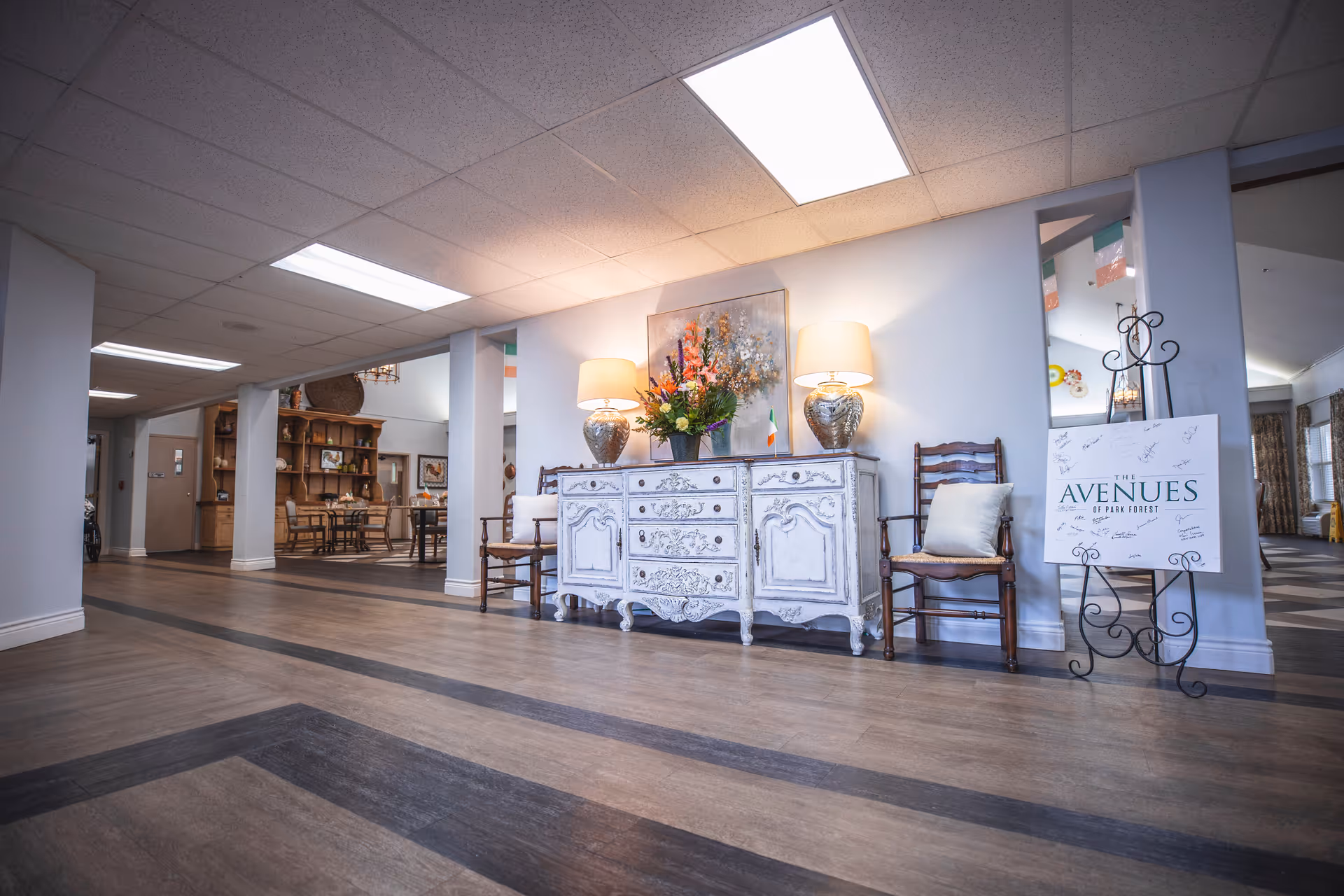 A spacious interior hallway with wooden flooring and white walls. There is a white ornate sideboard with two lamps and a flower arrangement on top, flanked by two wooden chairs with cushions. A large painting hangs above the sideboard. To the right, a sign on an easel reads 'The Avenues of Park Forest' with signatures. In the background, there are dining tables and chairs, and some decorative elements on the walls.