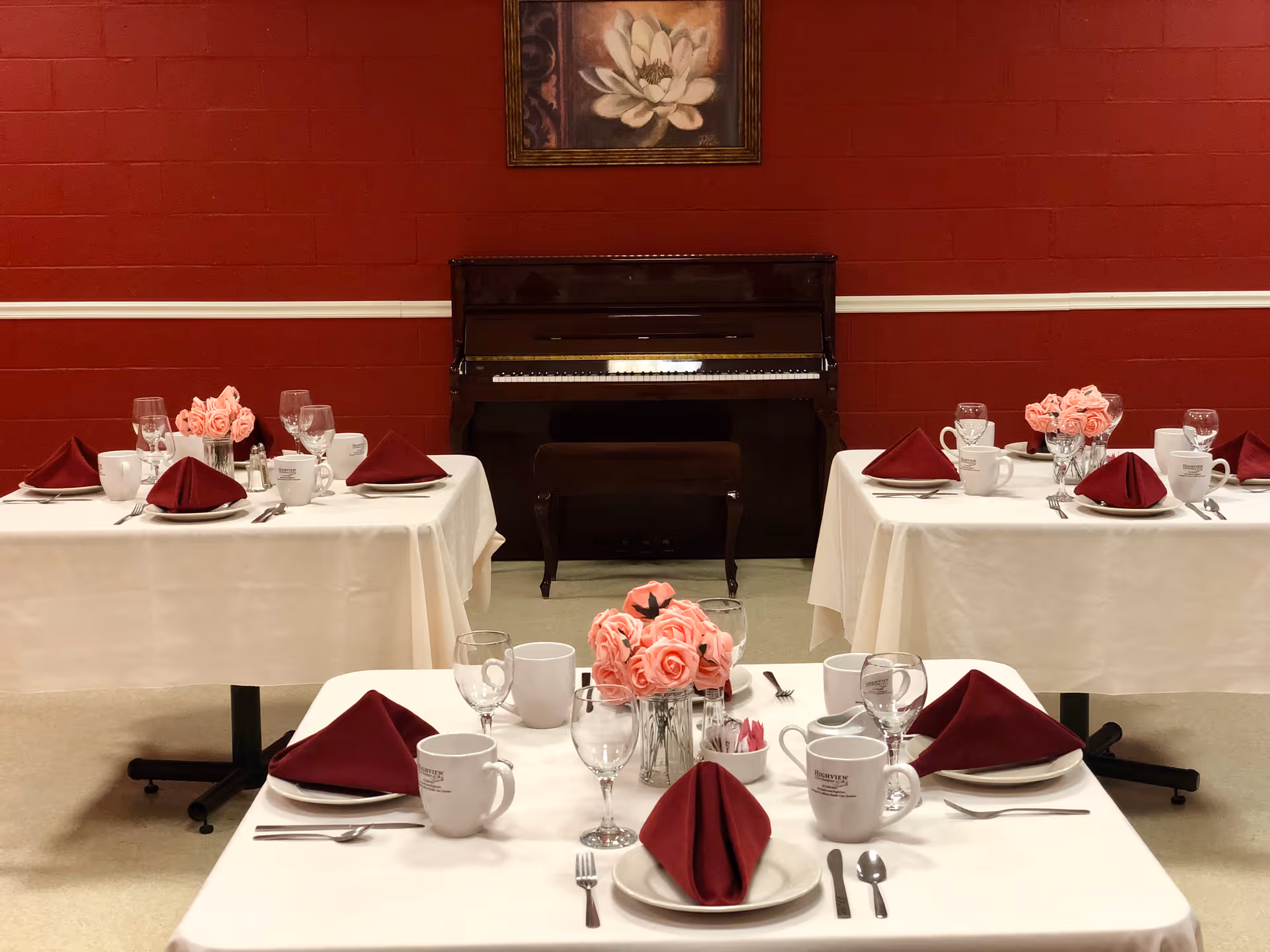 A dining room with three tables set for a meal. Each table is covered with a white tablecloth and has maroon folded napkins, white plates, silverware, glassware, and white mugs. Pink rose centerpieces are placed on each table. In the background, there is a dark brown upright piano against a red brick wall with a framed flower painting hanging above it.