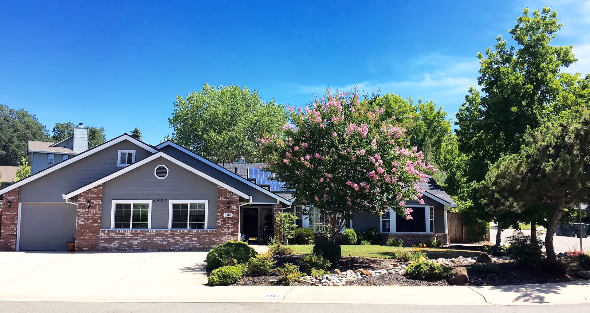 Exterior view of a single-story senior living facility building with a brick and gray siding facade, a garage on the left, and a well-maintained garden with green shrubs and a blooming tree with pink flowers in front. The sky is clear and blue.