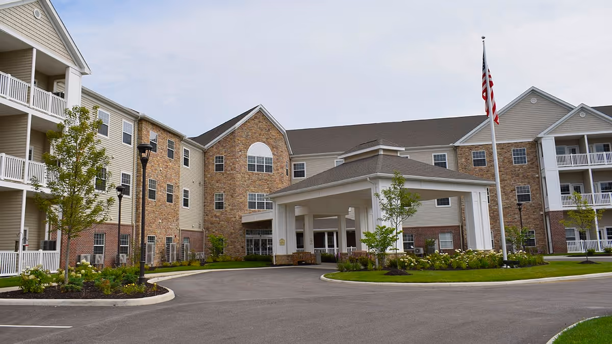 Exterior view of a multi-story senior living facility with a covered entrance, landscaped greenery, and an American flag on a flagpole in front.