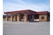 Exterior view of a single-story brick building with a flat roof and a covered entrance supported by columns, situated next to a large paved area under a clear sky.