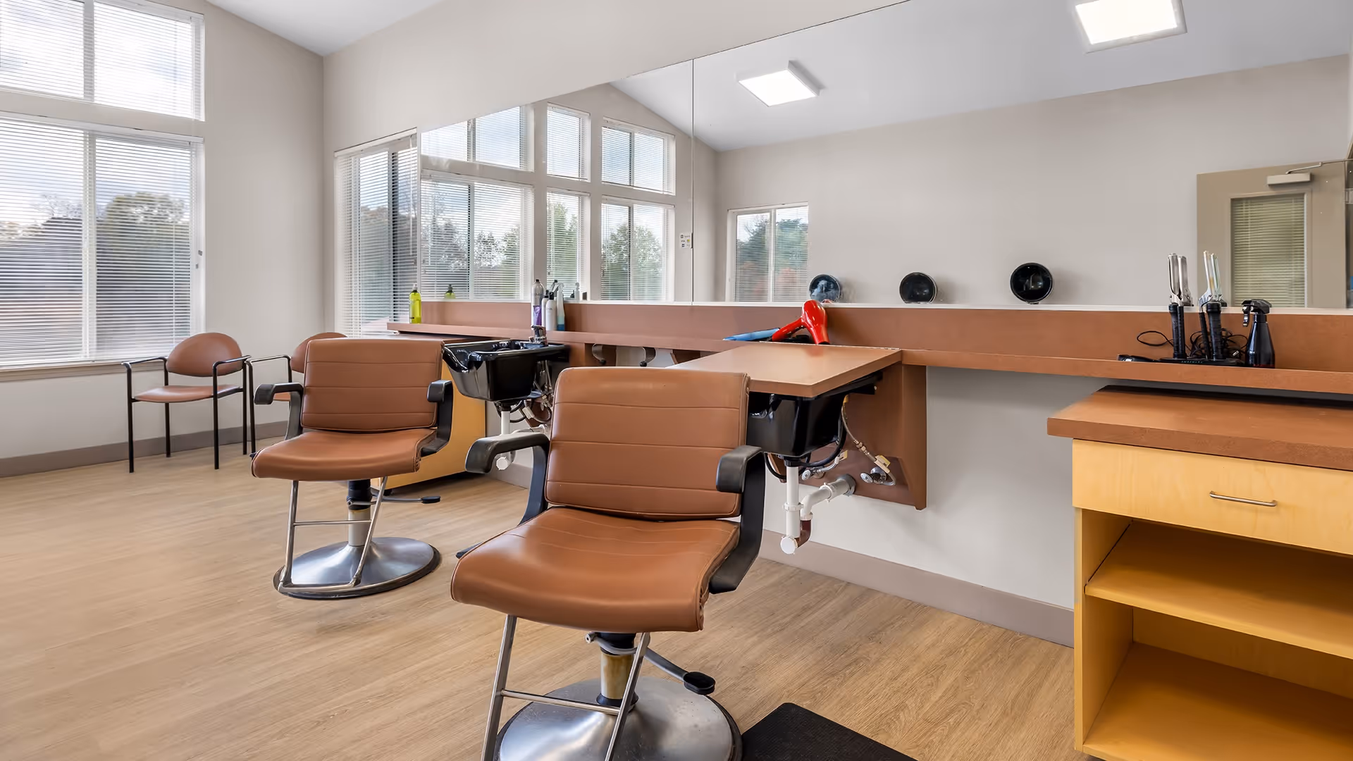Interior view of a hair salon area with two brown salon chairs in front of a long mirror. There are hair washing sinks attached to the counter, a hairdryer, and various hair care tools on the counter. Large windows with blinds allow natural light into the room.