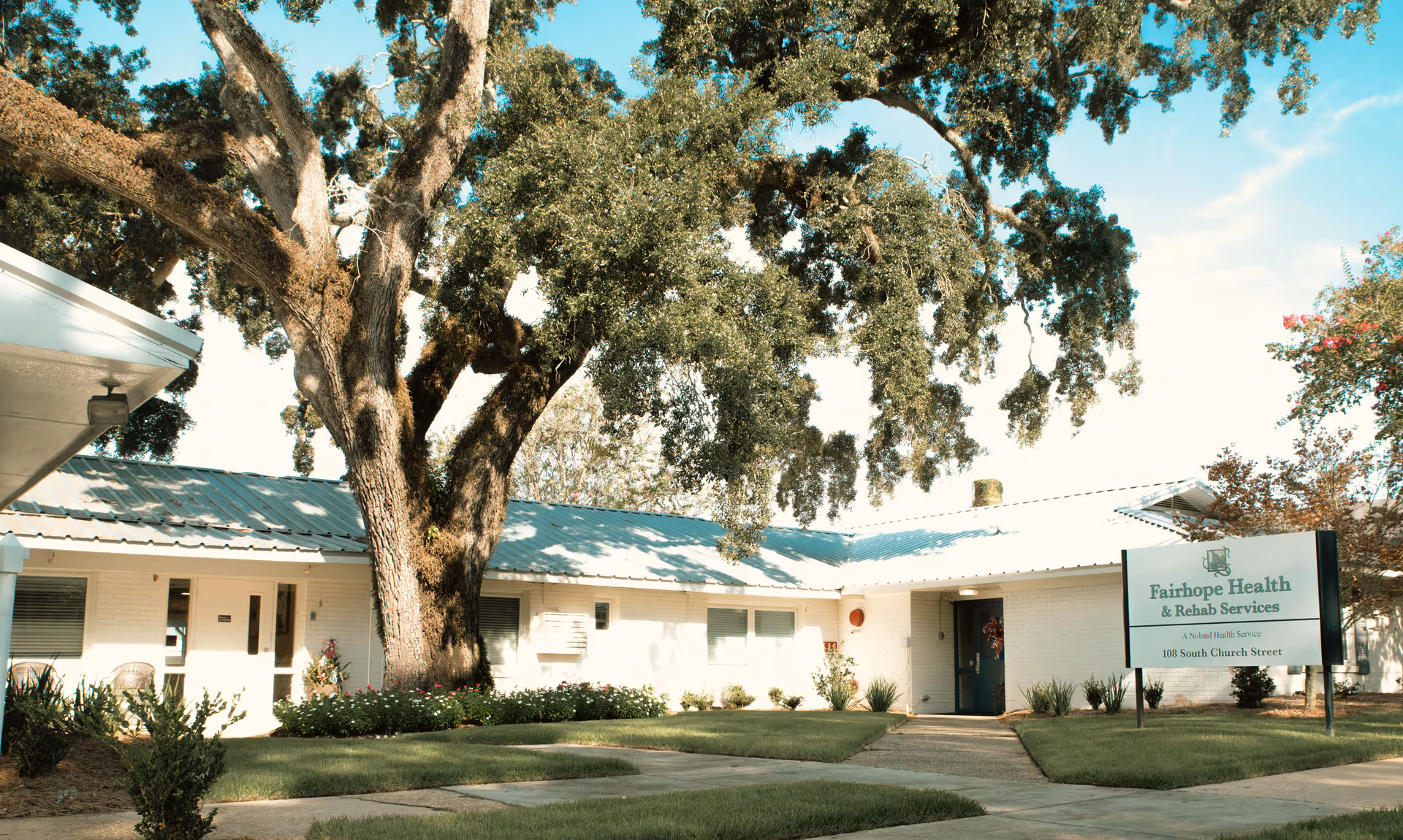 Exterior view of Fairhope Health & Rehab Services building with a large tree in front, well-maintained lawn, and a sign displaying the facility's name and address at 108 South Church Street.
