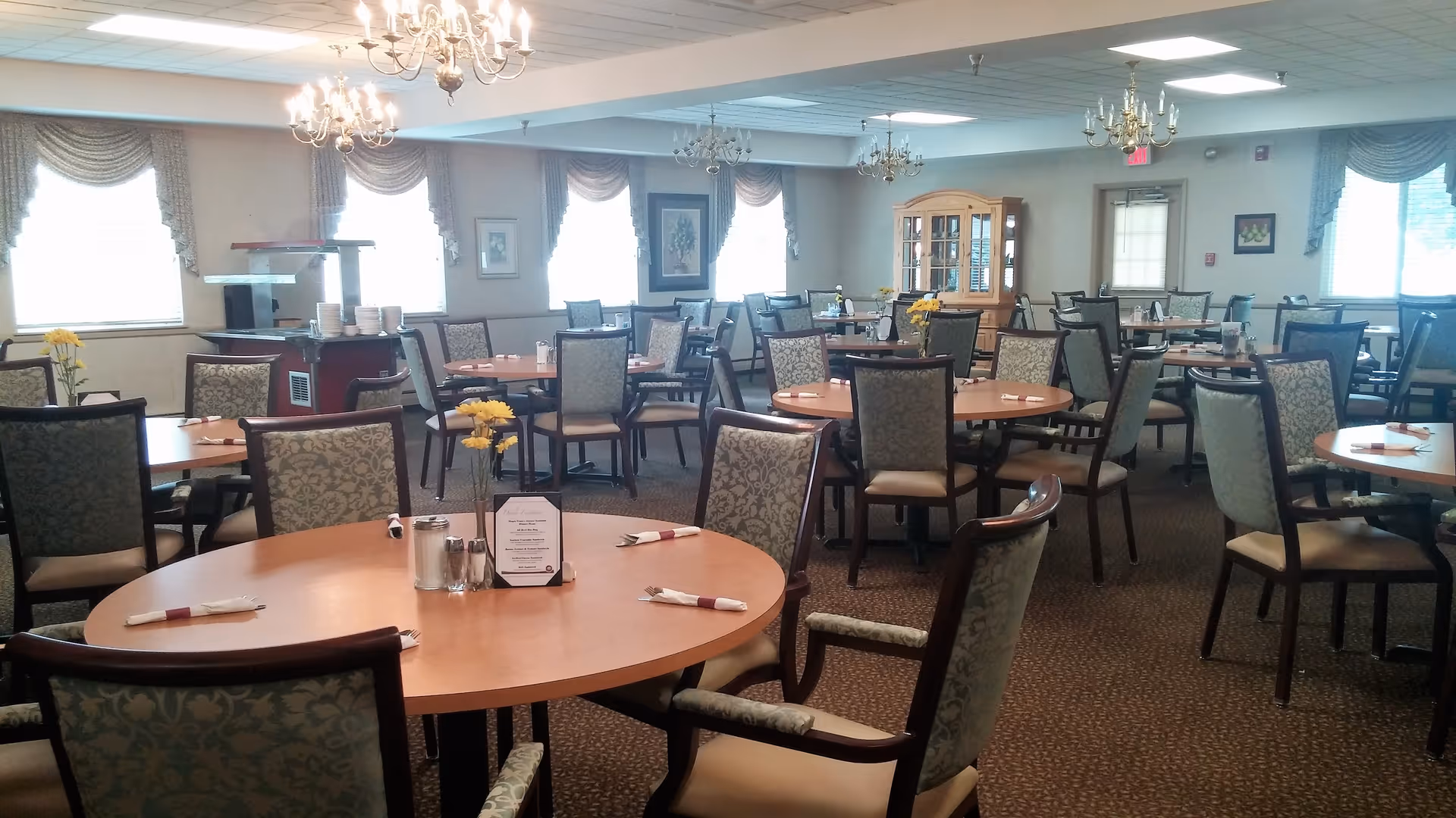 A spacious dining room with multiple round wooden tables and cushioned chairs arranged neatly. Each table has a small centerpiece with yellow flowers, salt and pepper shakers, and napkins wrapped around utensils. The room is well-lit with natural light coming through windows adorned with draped curtains. Chandeliers hang from the ceiling, and a wooden cabinet is visible against the far wall.