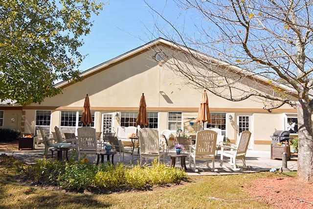 Outdoor patio area at a senior living facility with multiple beige chairs and tables arranged on a concrete surface. Several closed brown and orange umbrellas are positioned among the seating. The building behind the patio has beige walls and multiple windows and doors. Trees and shrubs surround the patio area under a clear blue sky.