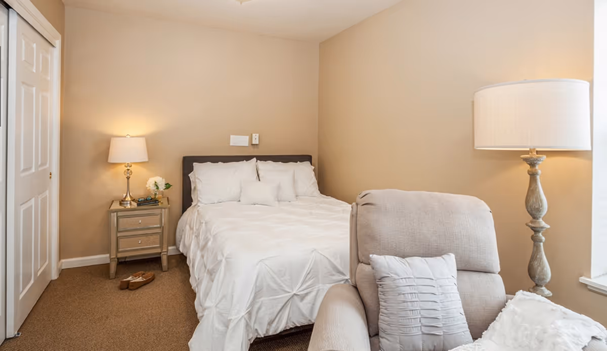 A neutral-toned bedroom with a bed dressed in white linens, a nightstand with a lamp, and a cushioned armchair.