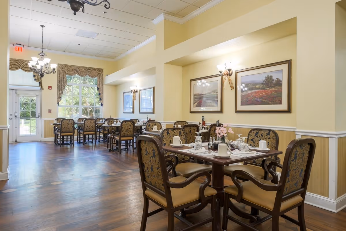 A dining room in a senior living facility with several tables and chairs arranged neatly. The tables are set with cups, glasses, and napkins. The room has wooden floors, yellow walls with white trim, framed landscape paintings, and wall sconces. Large windows with curtains allow natural light to enter, and a chandelier hangs from the ceiling.