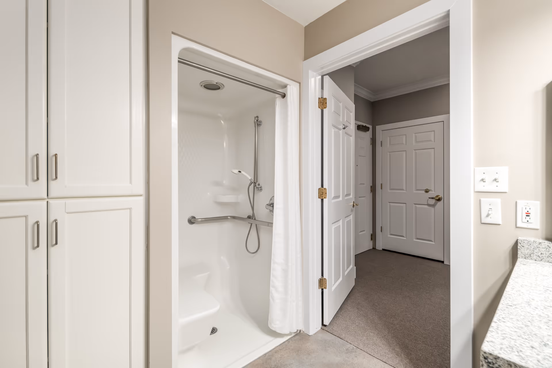 Accessible bathroom featuring a white walk-in shower with a grab bar and handheld shower head, adjacent cabinetry, and an open door to a carpeted hallway.