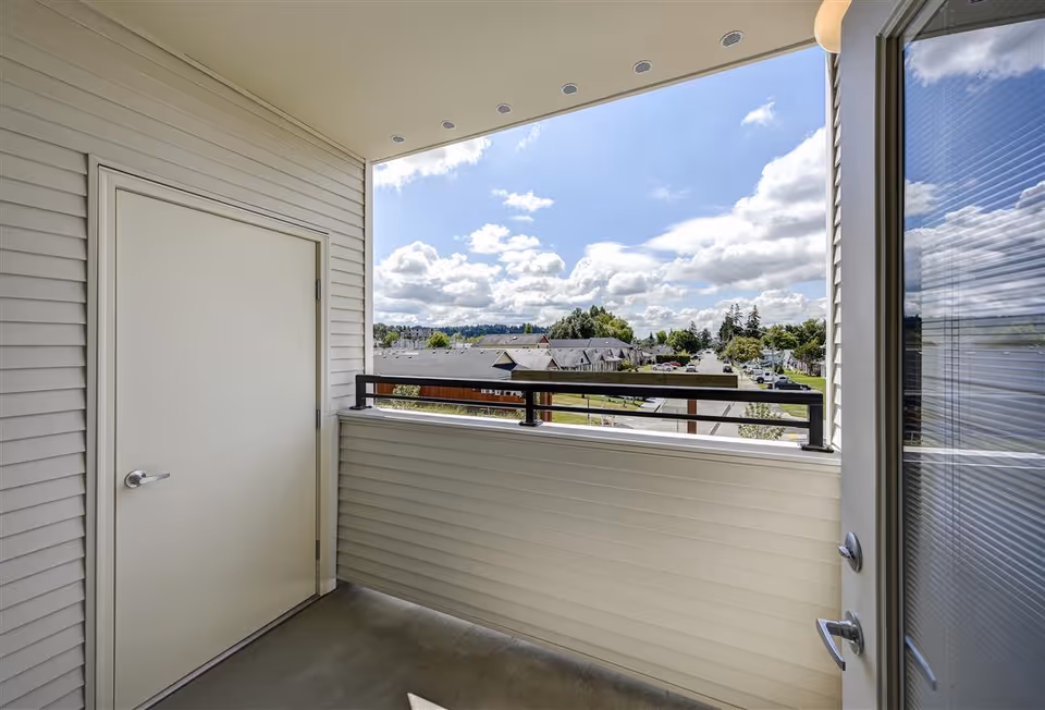 View from a covered balcony with beige siding and a closed door on the left. The balcony overlooks a neighborhood with houses, trees, and a partly cloudy sky.