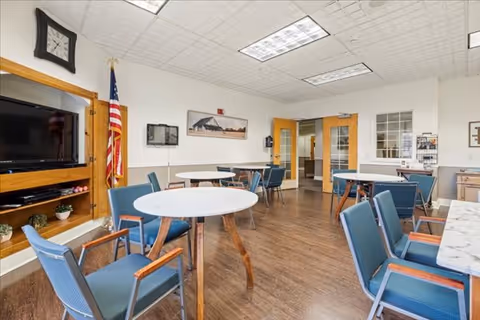 A bright common area with several round tables and blue cushioned chairs arranged around them. The room has wood flooring, a large flat-screen TV mounted on a wooden entertainment unit, an American flag in the corner, and a clock on the wall. There are double doors and a window leading to another room, and framed pictures hang on the walls.