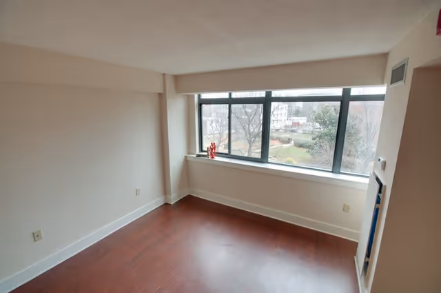 Empty room with wooden flooring, beige walls, a large window overlooking trees and a garden area outside, and a small red decorative item on the window sill.