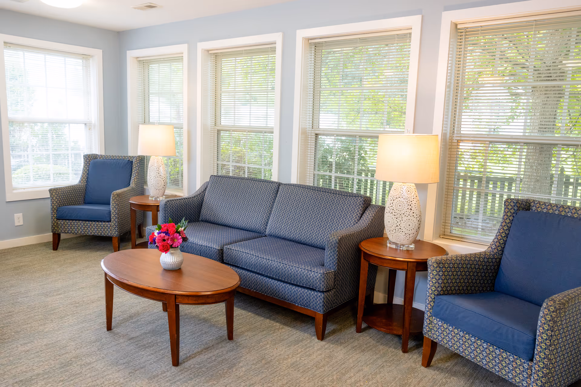A bright sitting area with a blue upholstered sofa and two matching armchairs around a wooden coffee table and side tables with lamps in front of large windows.