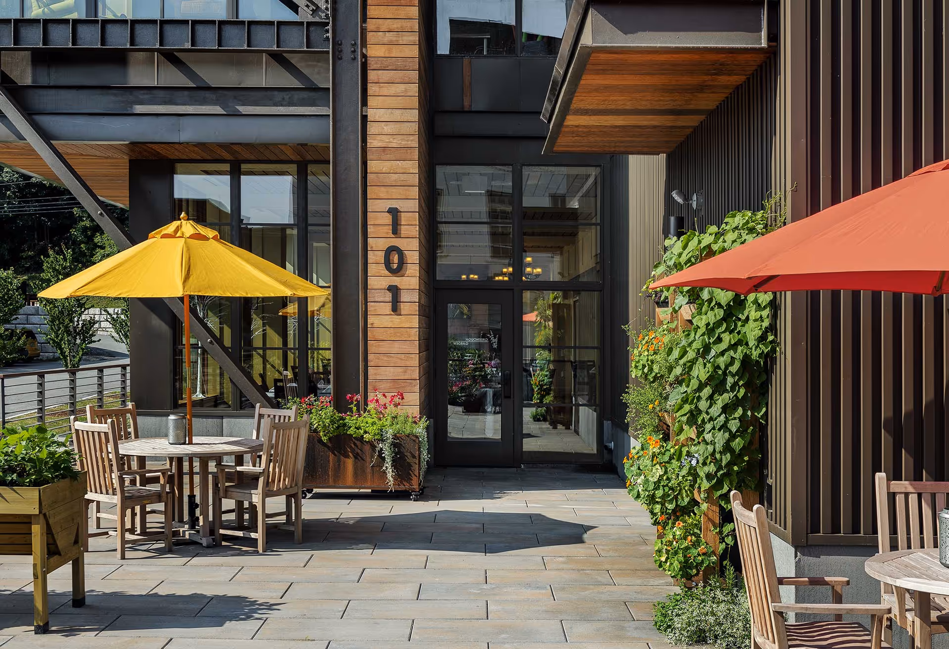 Outdoor patio area with wooden tables and chairs under yellow and red umbrellas, surrounded by planters with green plants and flowers, adjacent to a modern building entrance with large glass doors and the number 101 displayed on a wooden panel.