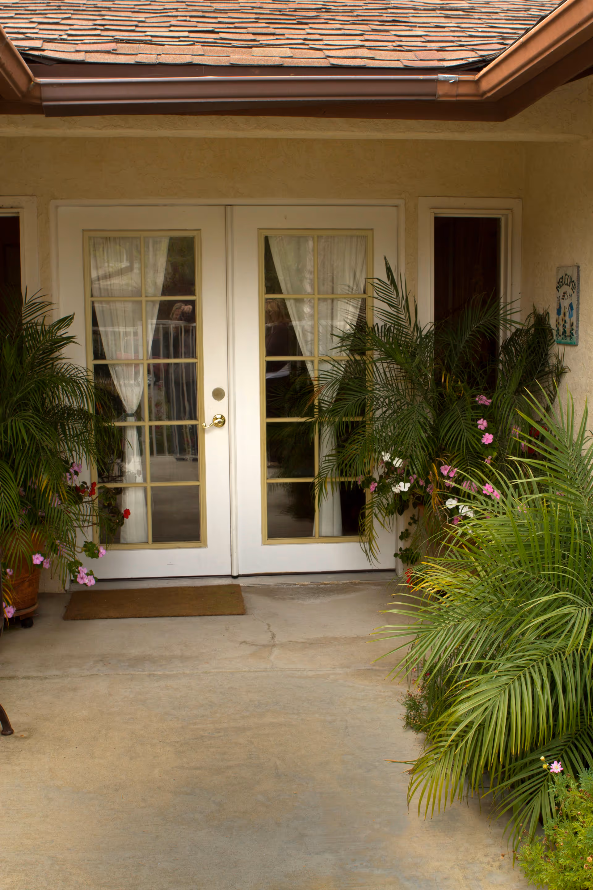 Entrance to a building with white double French doors featuring glass panes and white curtains. The doorway is flanked by lush green potted plants with some pink and white flowers. A brown doormat is placed in front of the doors on a concrete floor. The building has a beige exterior wall and a brown shingled roof.