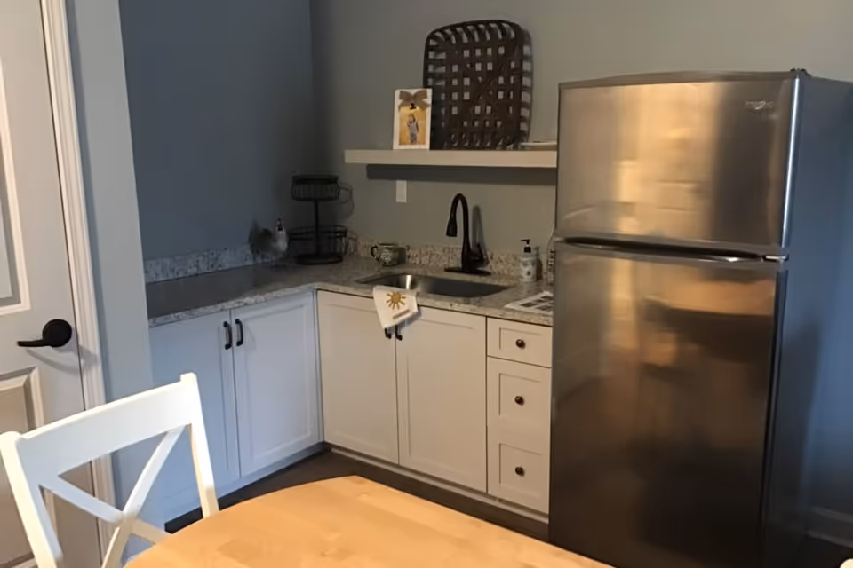 A small kitchen area with white cabinets, a granite countertop, a stainless steel refrigerator, a black faucet over a small sink, and a wooden table with a white chair in the foreground. There is a decorative basket and a framed picture on a floating shelf above the countertop.