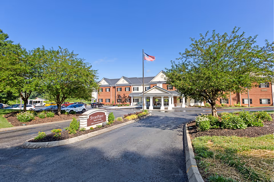 Exterior view of StoryPoint Knoxville West senior living facility showing a brick building with white trim, a covered entrance, an American flag on a flagpole, landscaped greenery, trees, and a parking area with several cars under a clear blue sky.