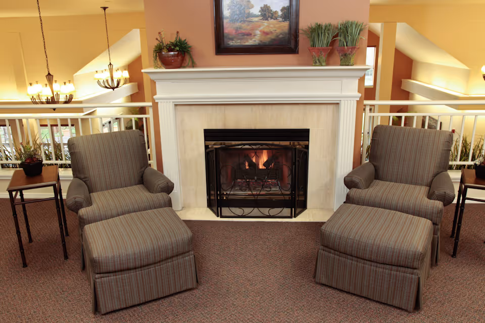 Two striped upholstered armchairs with matching ottomans are positioned in front of a lit fireplace with a white mantel. On the mantel are decorative plants and a framed landscape painting. Small side tables with potted plants flank each chair. The room has warm lighting and a carpeted floor.