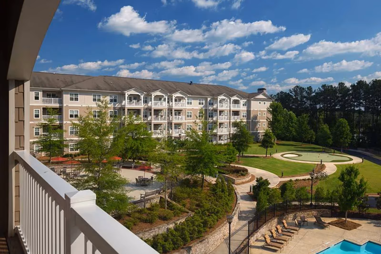 View from a balcony overlooking a landscaped courtyard with a pool and a multi-story senior living building under a blue sky.