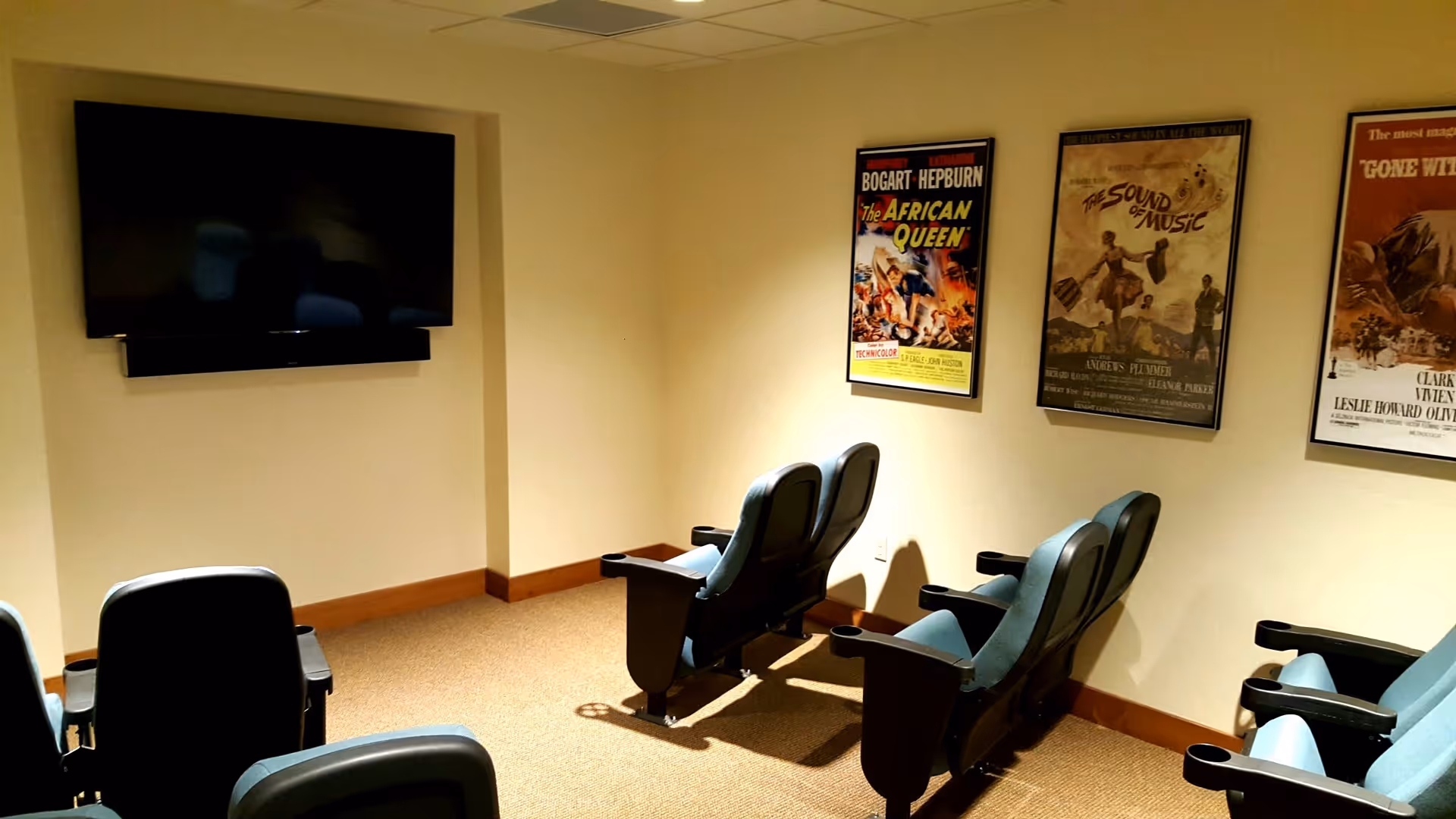 Small theater room with six theater-style seats arranged in two rows facing a large flat-screen TV mounted on the wall. Three framed classic movie posters are displayed on the adjacent wall.