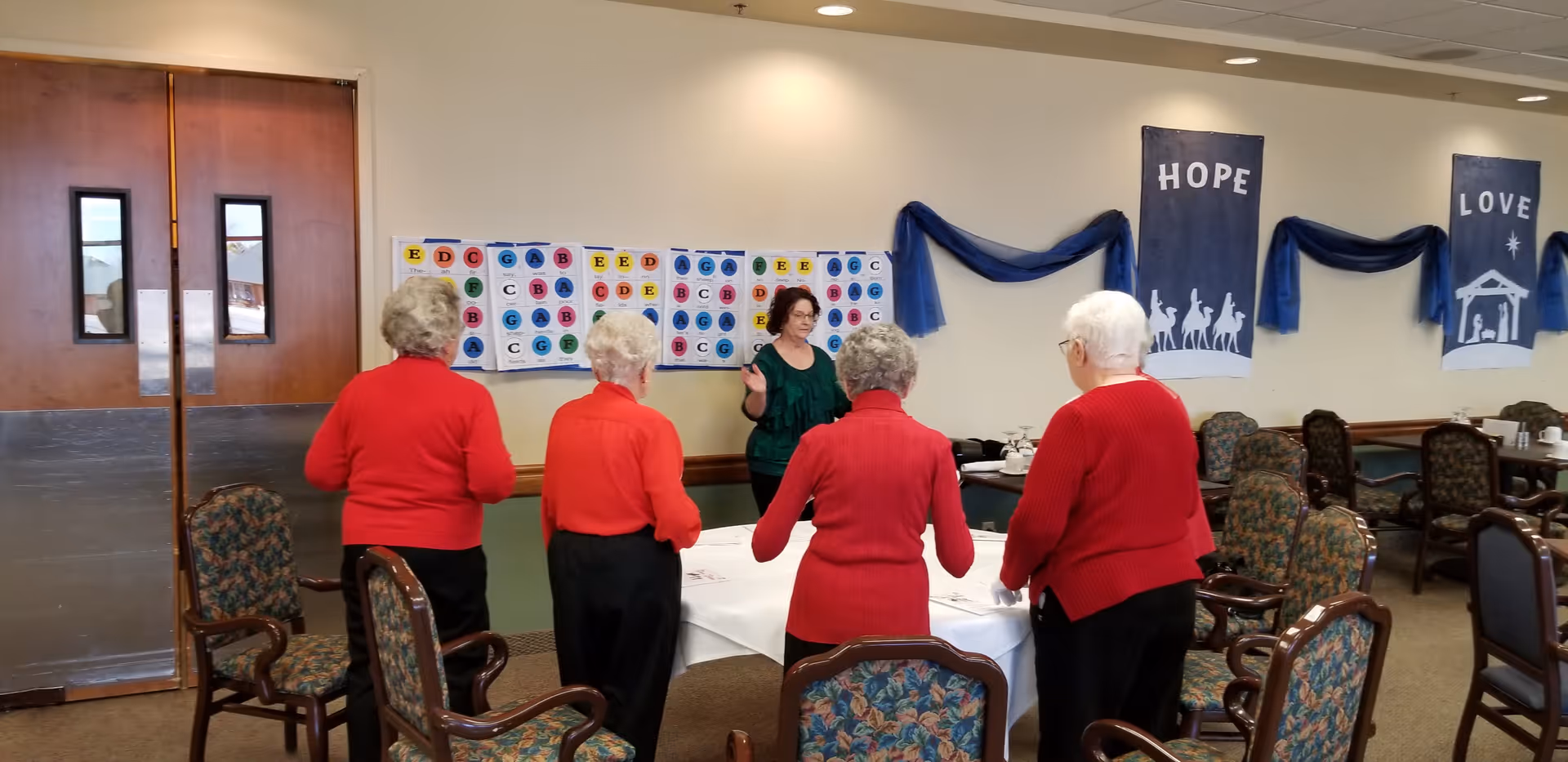Several senior women in red sweaters stand around a table in a communal dining/activity room with banners reading 'HOPE' and 'LOVE' on the wall.