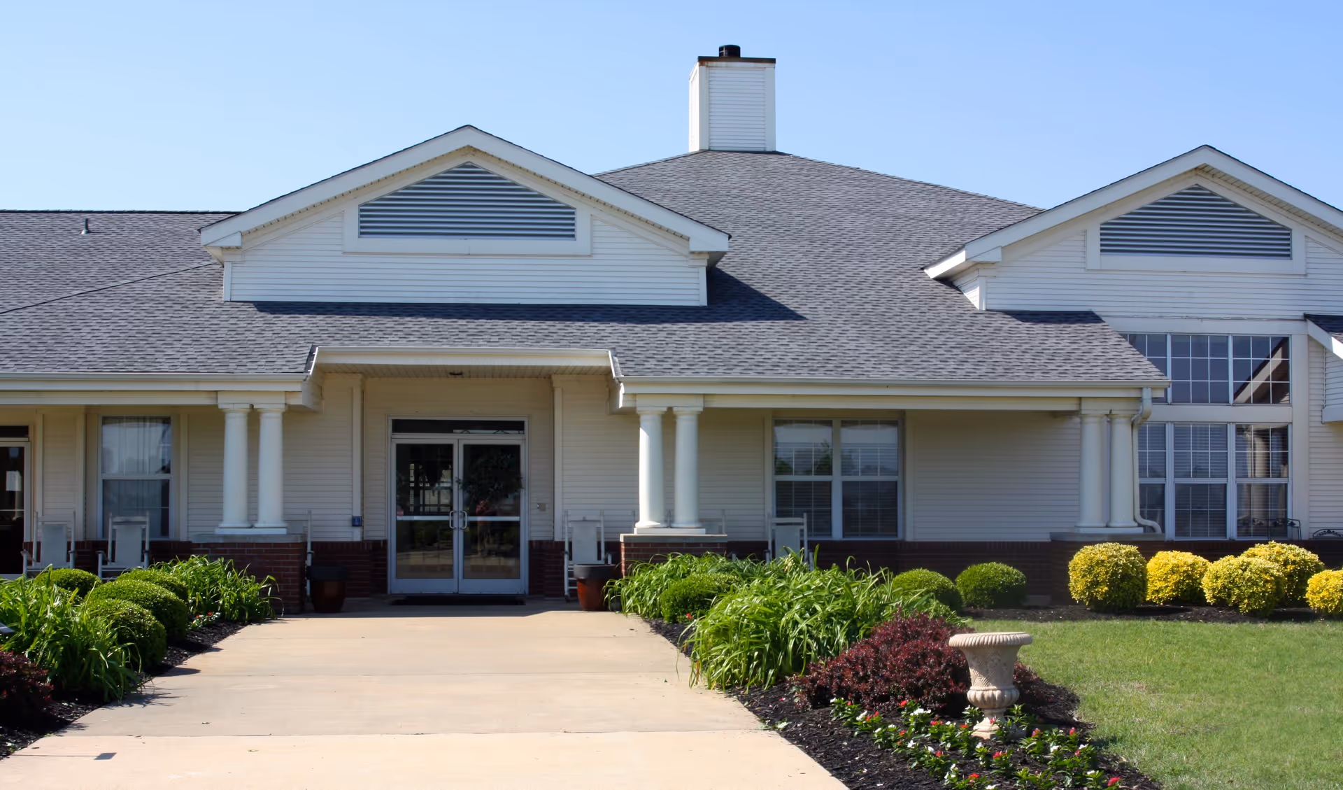 Front entrance of a single-story senior living building with columns, a landscaped walkway, and double glass doors.