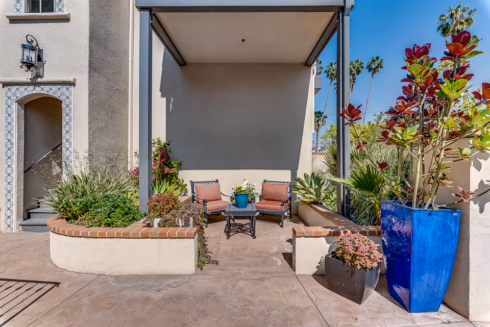 Outdoor seating area with two cushioned chairs and a small table with a potted plant, surrounded by various plants and flowers in planters, next to a building with a tiled archway and stairs.