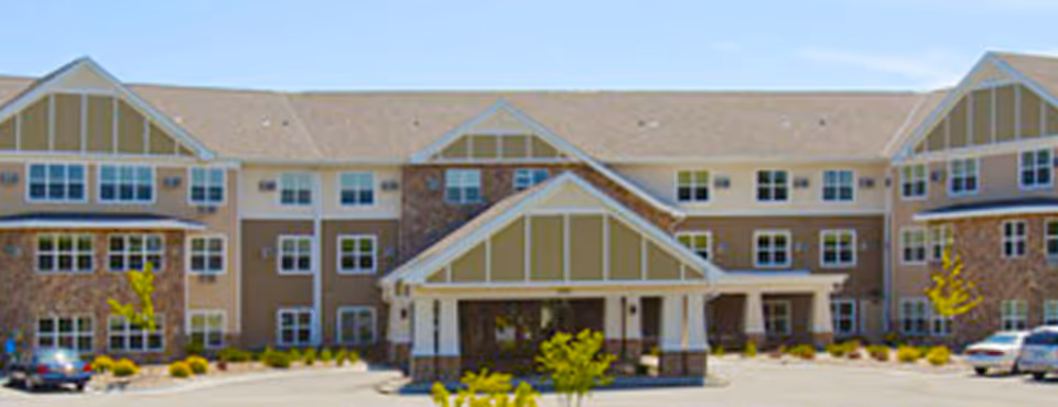 Front exterior view of a large multi-story senior living facility building with beige and brown walls, multiple windows, and a covered entrance. There are a few cars parked in front and some small trees and shrubs in the landscaping.