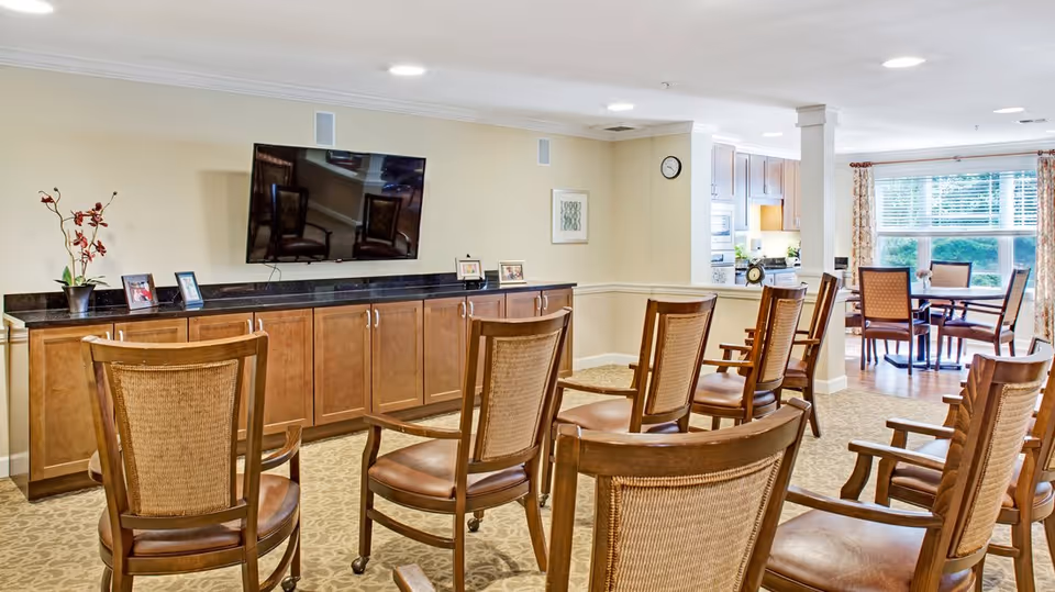 Bright communal lounge with wooden chairs arranged facing a wall-mounted TV and cabinetry, and a dining area visible by large windows.