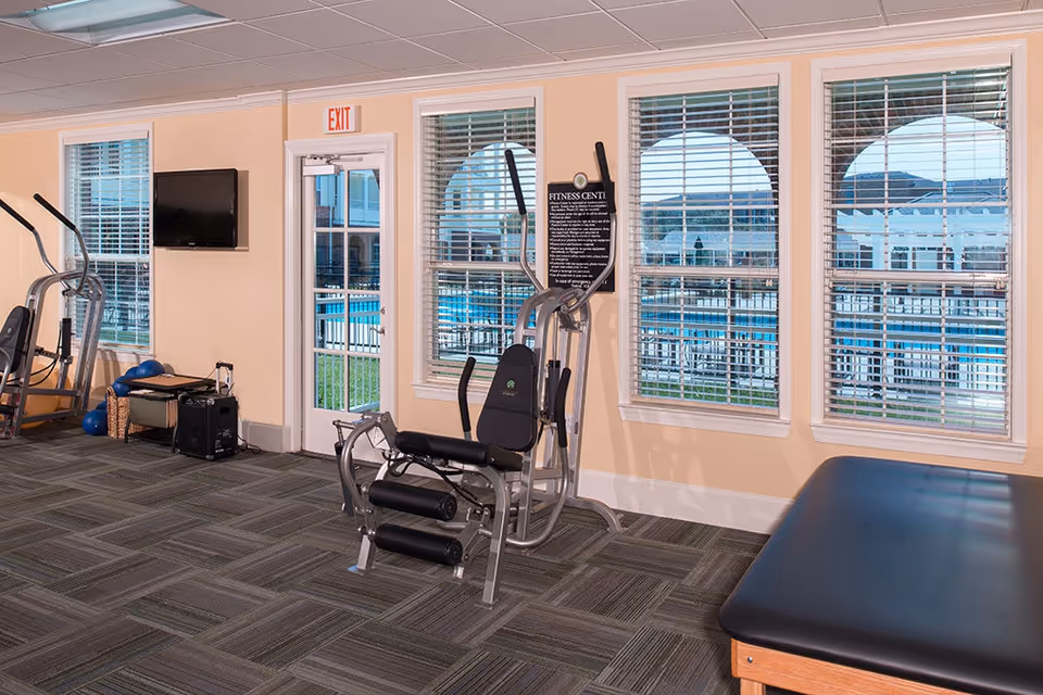 Exercise room with a weight machine, cardio equipment, and a treatment table, with windows overlooking an outdoor pool and an exit door.