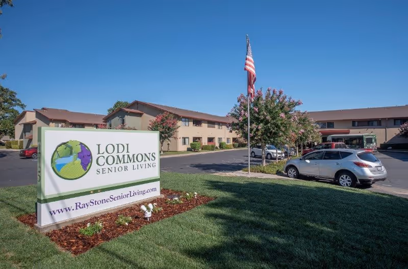 Exterior view of Lodi Commons Senior Living facility with a large sign in the foreground displaying the facility name and website. The building is two stories with beige walls and brown roofs. There is a parking lot with several cars and an American flag on a flagpole near the center of the image. Trees with pink flowers are visible around the parking area under a clear blue sky.