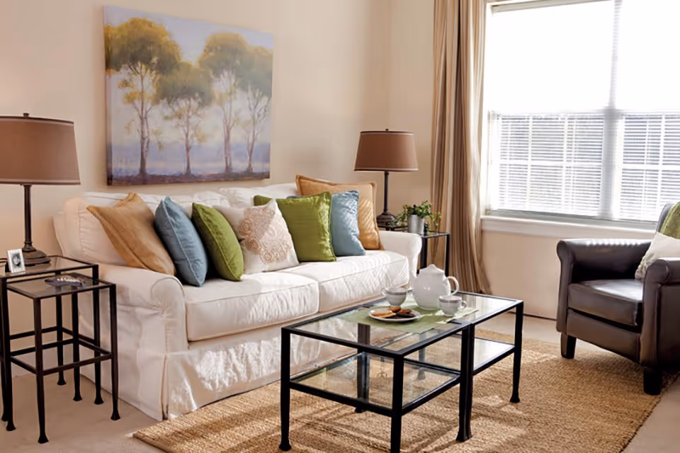 Sunlit living room with a white sofa topped with colorful throw pillows, a glass coffee table, armchair and table lamps.