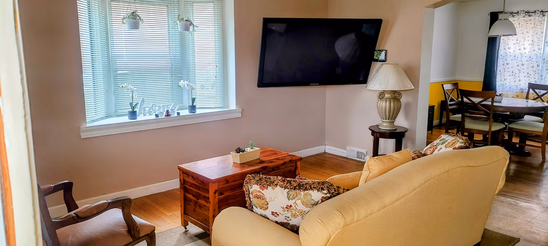 A cozy living room with a beige sofa adorned with floral cushions, a wooden coffee table, and a wooden armchair. A flat-screen TV is mounted on the wall. There is a window with blinds and small potted plants on the windowsill. In the background, a dining area with a round table and chairs is visible.