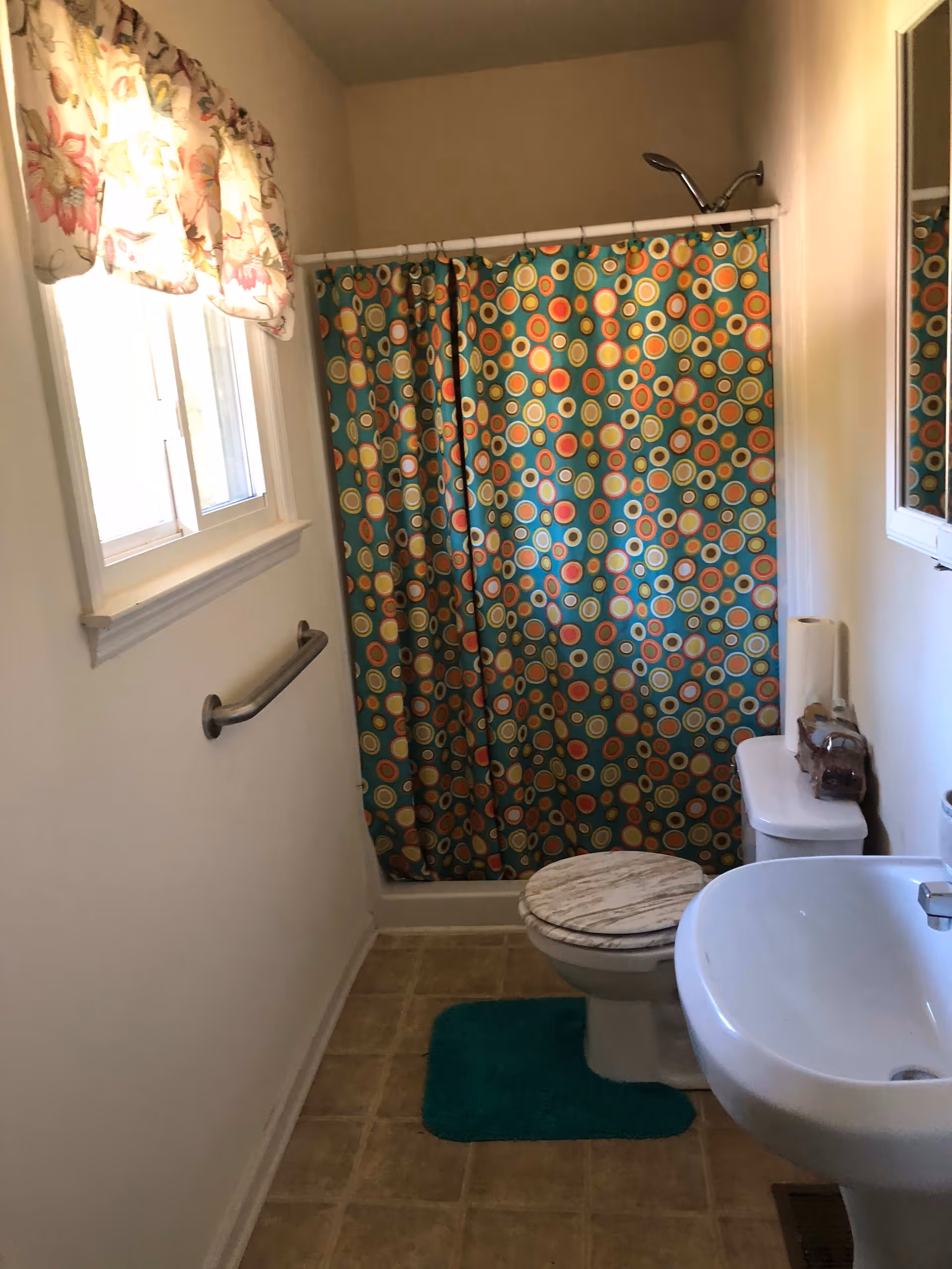 Small bathroom with a colorful circular-patterned shower curtain, pedestal sink, toilet with a marble-patterned seat, window with floral valance and a wall grab bar.
