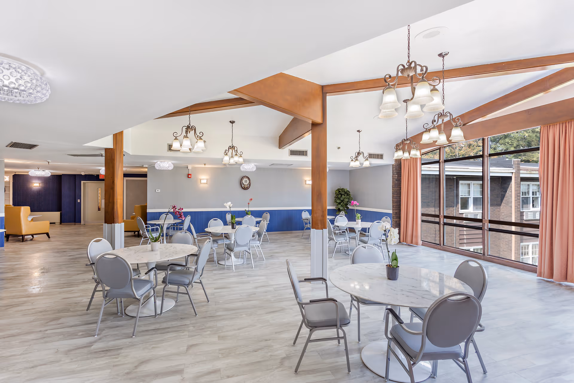 A bright and spacious dining area with multiple round tables and gray chairs arranged neatly. The room features large windows with peach-colored curtains allowing natural light to fill the space. Wooden beams and multiple hanging light fixtures add to the decor. There are small potted plants on the tables and a clock on the far wall.