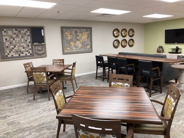 A communal dining or lounge area with several wooden tables and chairs featuring floral upholstery. The room has a bar counter with high stools, a wall-mounted TV, decorative wall art including a world map and circular plates, and a neutral color scheme with light green and beige walls.