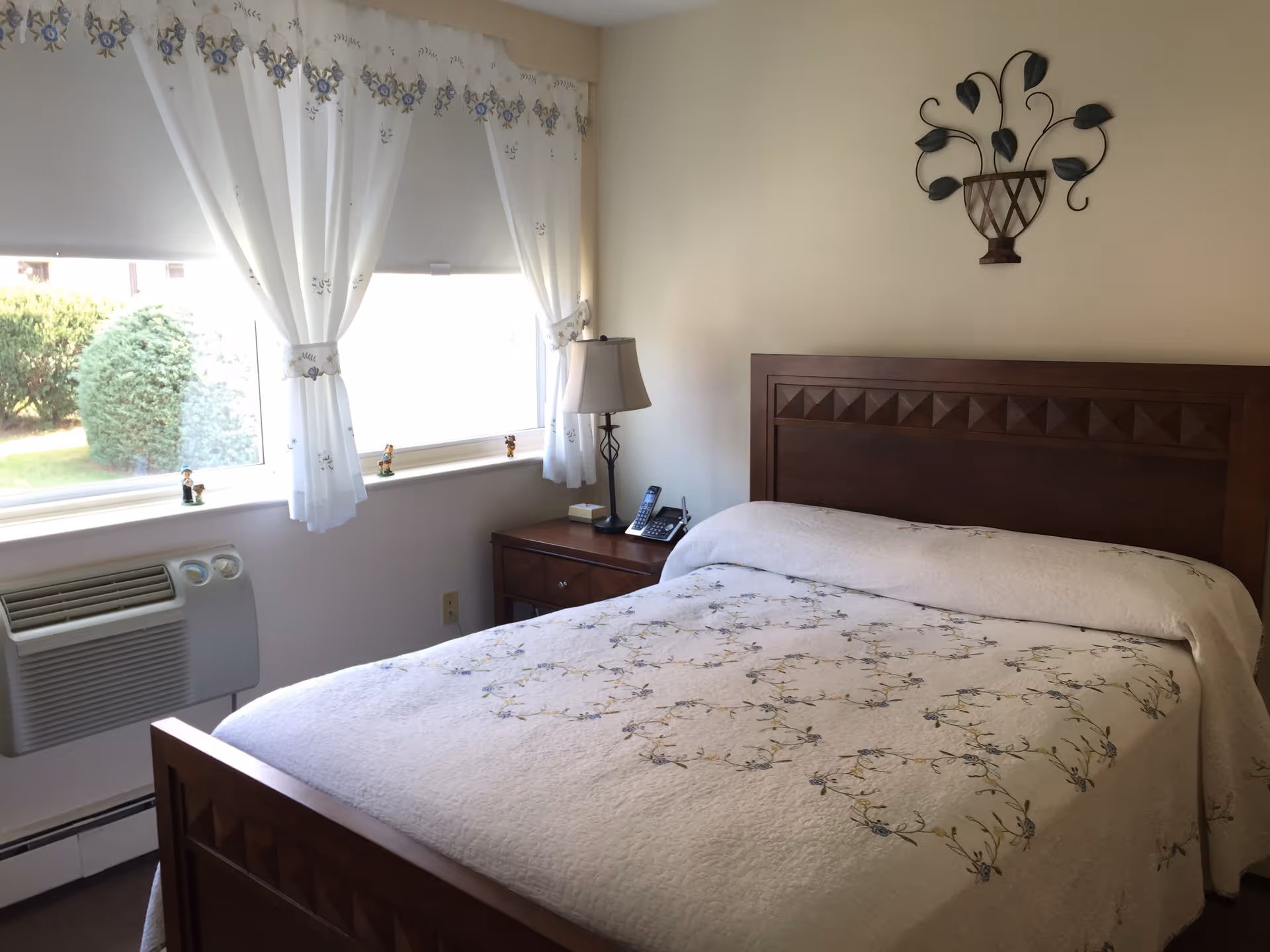 Sunlit bedroom with a made double bed featuring a wooden headboard, a nightstand with a lamp and phone, and a window with white curtains showing shrubs outside.