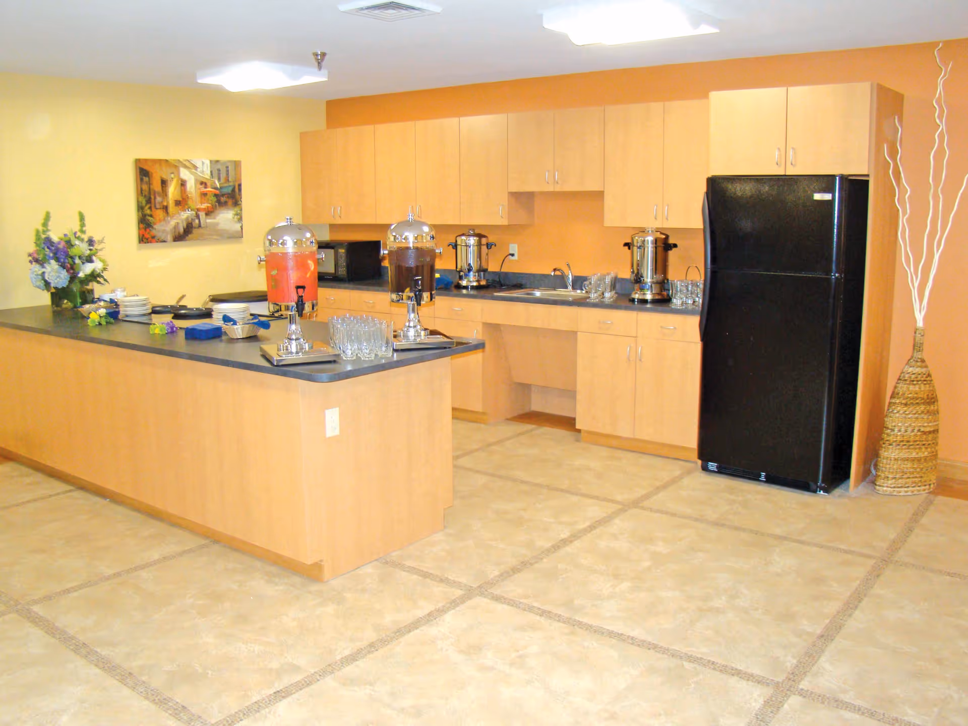 Communal kitchen area with a long island, beverage dispensers, light wood cabinets and a black refrigerator.