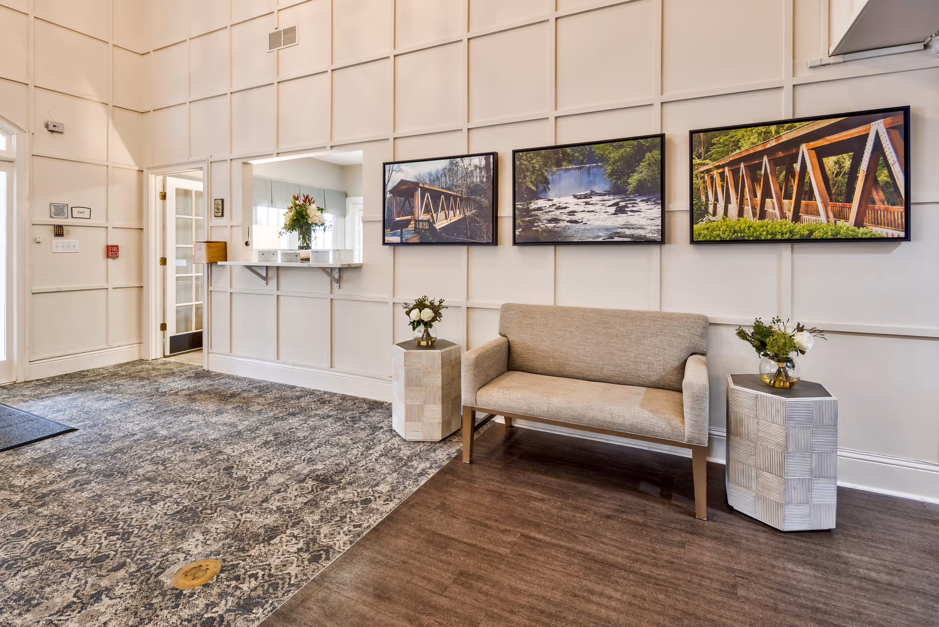 A bright and welcoming interior seating area with a beige loveseat flanked by two small tables holding flower arrangements. The wall behind features three framed landscape photographs of bridges and a waterfall. To the left, there is a doorway with glass-paneled double doors and a pass-through window with a flower vase on the counter. The floor transitions from patterned carpet to wood flooring.