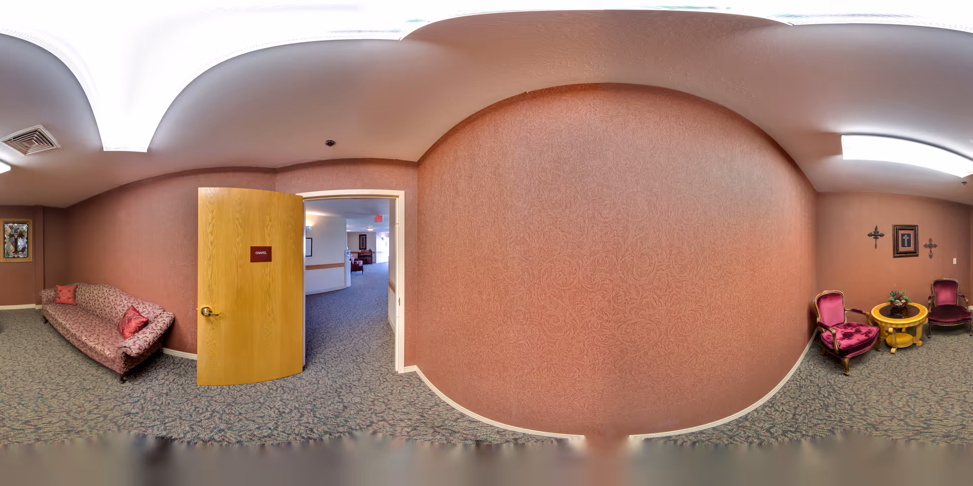 Carpeted interior lounge area with rose-patterned walls, a sofa on the left, an open wooden door leading to a corridor, and two upholstered chairs with a round table on the right.