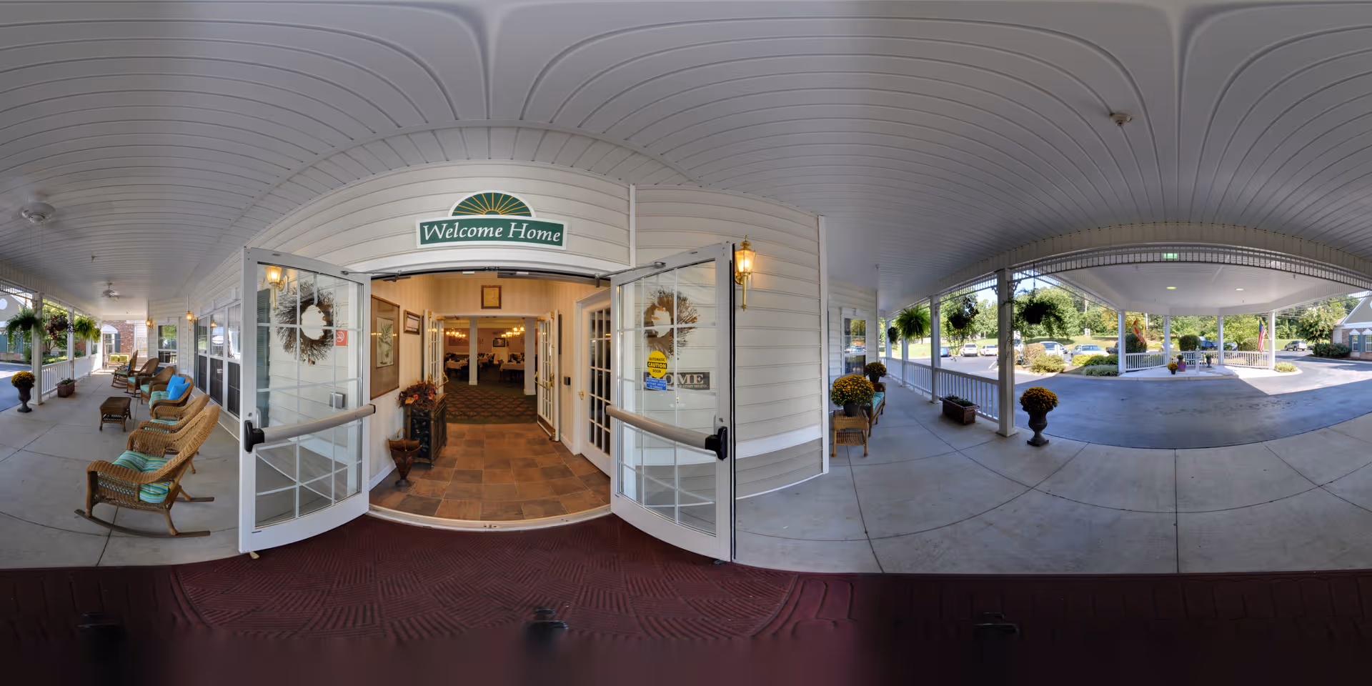 Covered entrance and porch of a senior living facility with open double doors under a 'Welcome Home' sign, wicker chairs, and a drive-through porte-cochere.