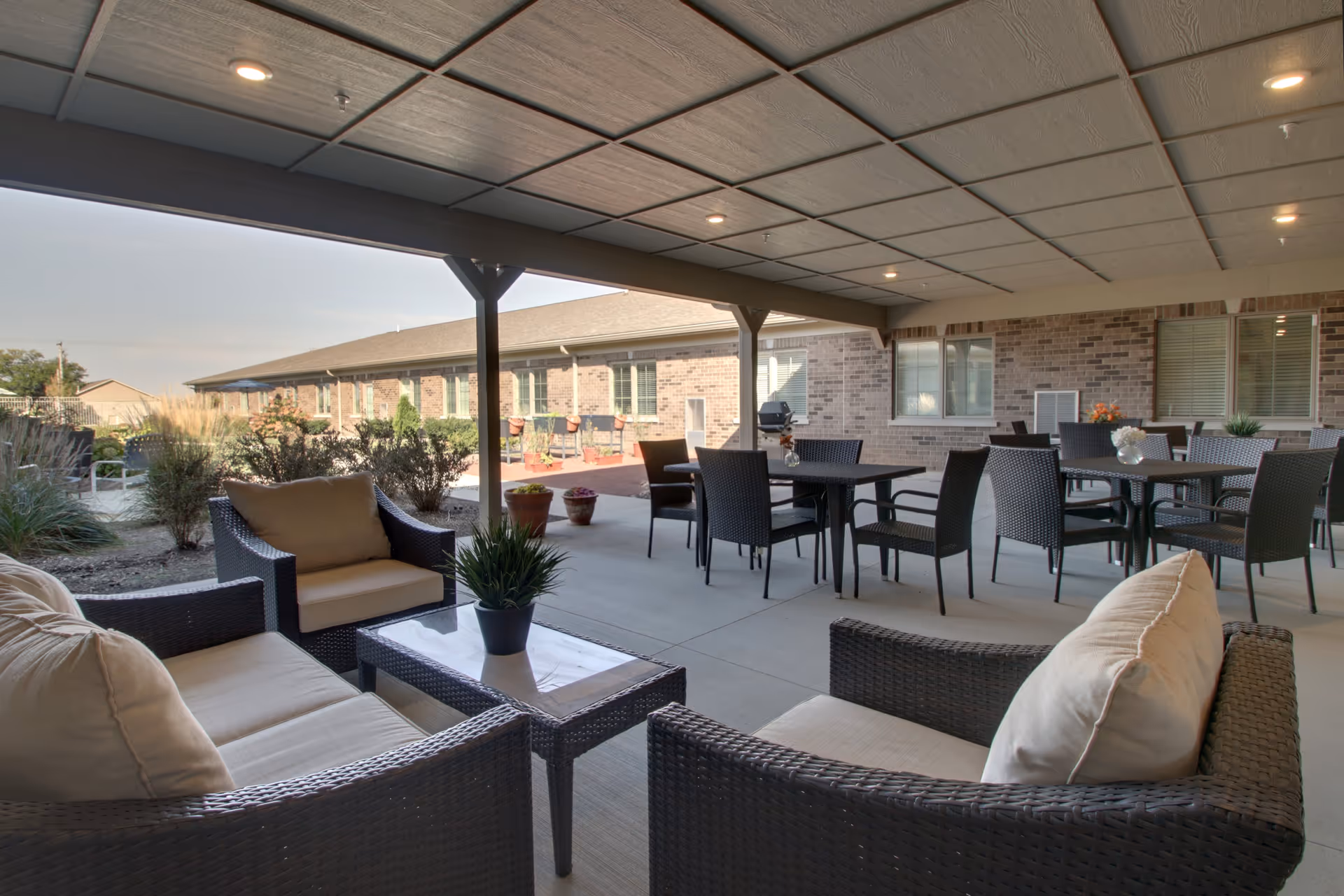 Covered outdoor patio area with wicker seating including cushioned chairs and sofas, several tables with chairs, potted plants, and a brick building in the background under a cloudy sky.