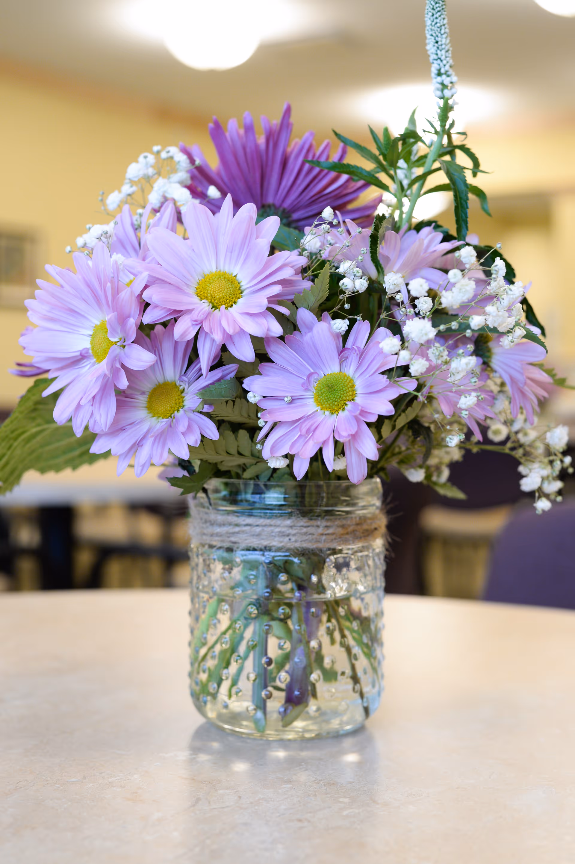 A bouquet of light purple daisies and white baby's breath flowers arranged in a clear glass jar with decorative dots and twine wrapped around the neck, placed on a beige table with a blurred indoor background.