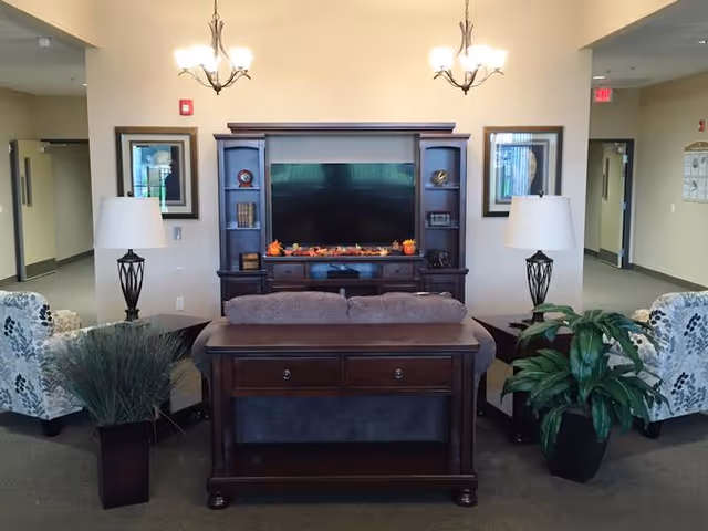 A cozy living room area with a large flat-screen TV mounted on a dark wooden entertainment center decorated with small autumn-themed items. In front of the TV is a gray sofa with a wooden console table behind it. Two patterned armchairs flank the sofa, each next to a table lamp with a white shade. There are two potted plants on either side of the seating area. The room has beige walls and carpeted floors, with two chandeliers hanging from the ceiling and framed artwork on the walls.