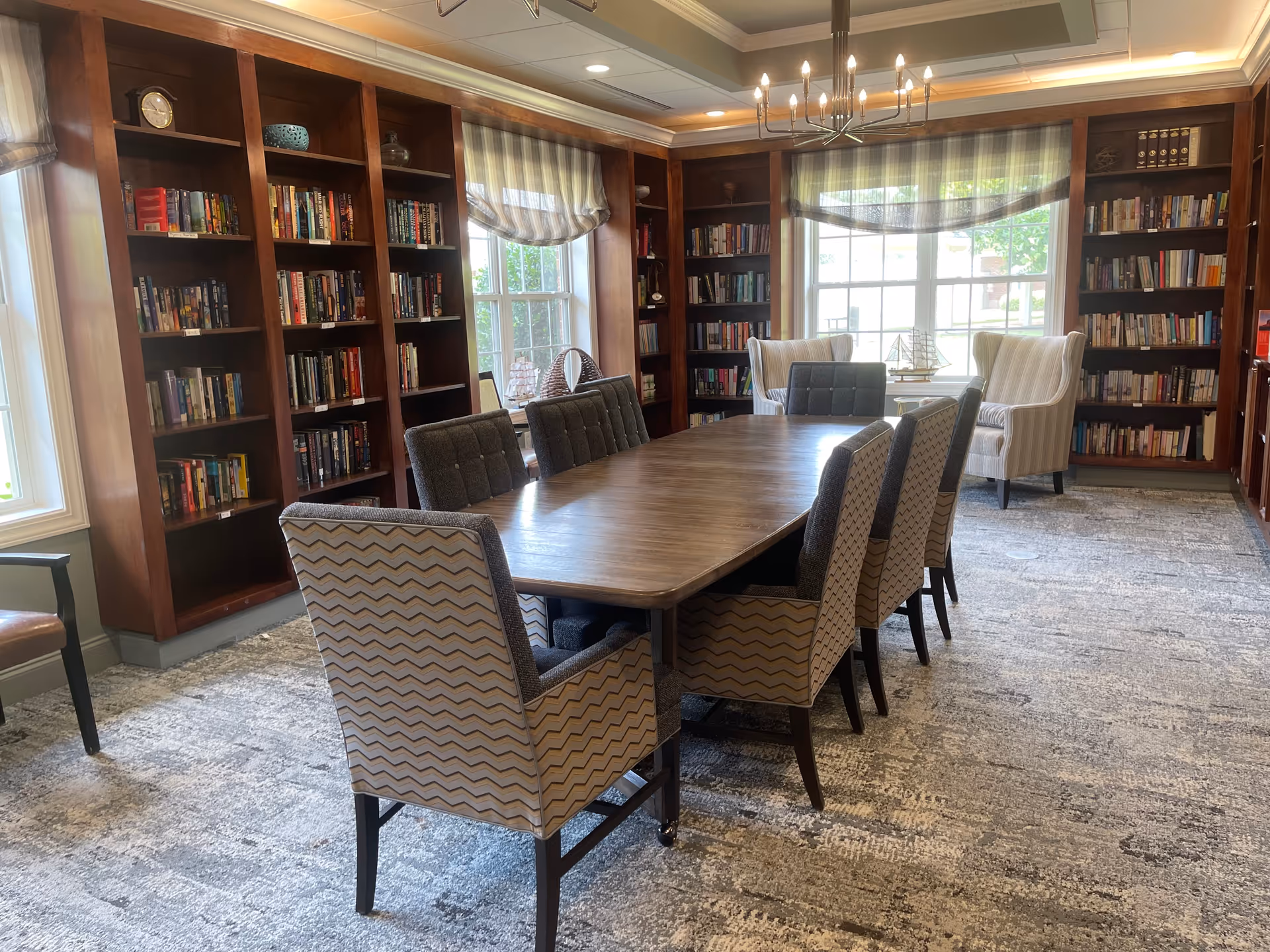 A cozy library room with wooden bookshelves filled with books along the walls, a large wooden table surrounded by eight upholstered chairs, two armchairs near the window, and a chandelier hanging from the ceiling. The room has large windows with striped curtains allowing natural light to enter.