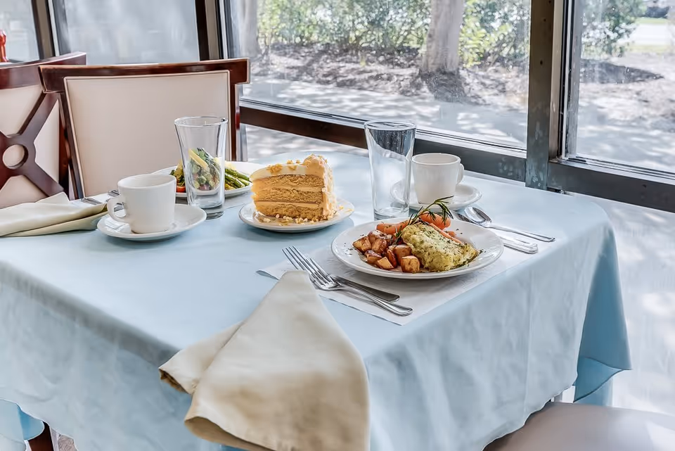 A dining table by a window set with plates of food including a slice of cake, a savory entree, coffee cups, glasses, and napkins.